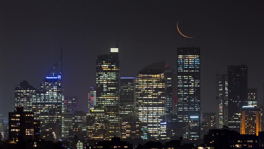 Crescent moon setting behind buildings on the city skyline with glowing lights