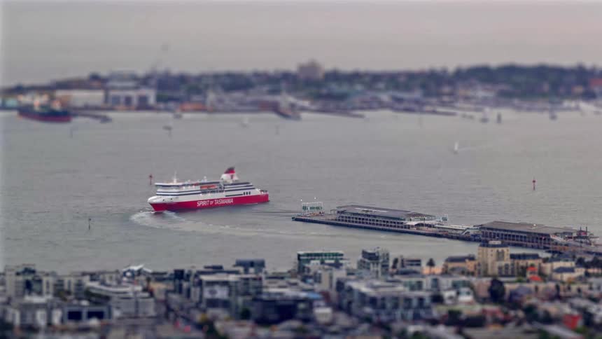 A view from a distance shows a large red and white ship moving through a body of water near a city during the day