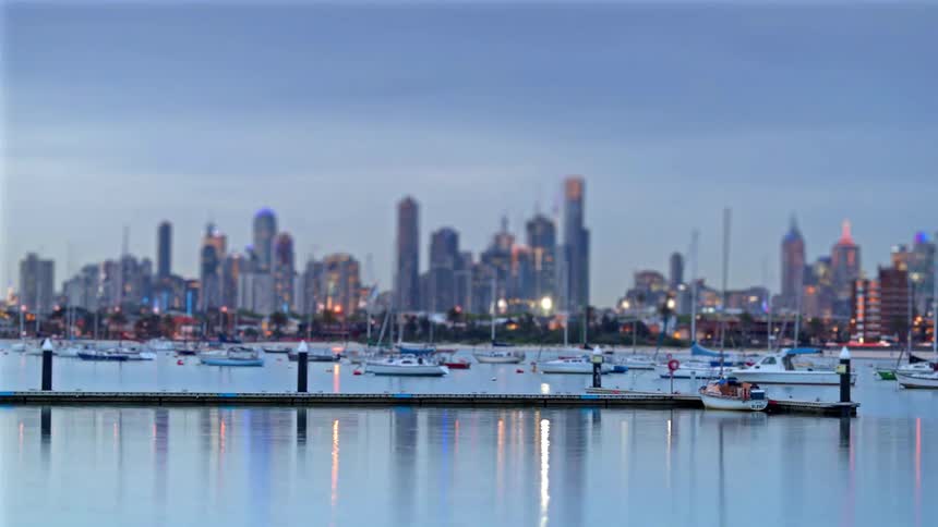 A harbor filled with boats transitions from sunset into a glittering nightscape against a distant city skyline