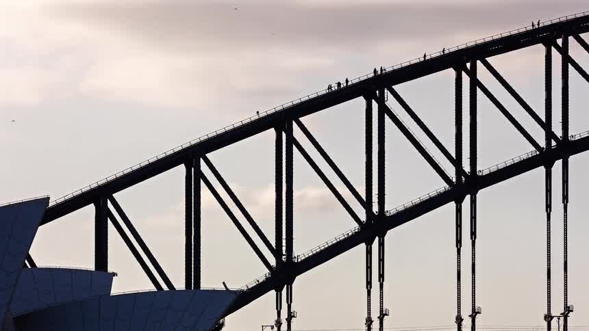 Steel arch bridge structure with pedestrians visible on the walkway against a cloudy sky