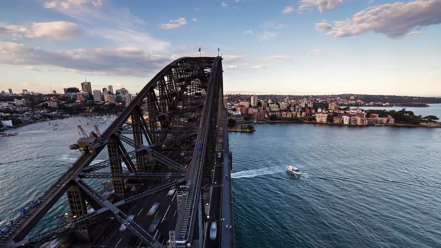 A highangle timelapse captures the iconic sydney harbour bridge transitioning from bright daylight to a vibrant night as heavy traffic flows along the deck tiny climbers scale the upper beams and ferries weave across the shimmering waters of sydney ha
