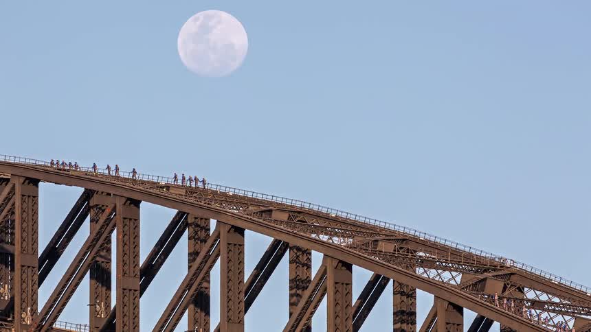 A large group of people walking on top of a steel arch bridge at sunrise with the moon still visible in the sky