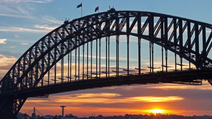 The sydney harbour bridge is silhouetted against a glowing sunset as clouds drift across the sky