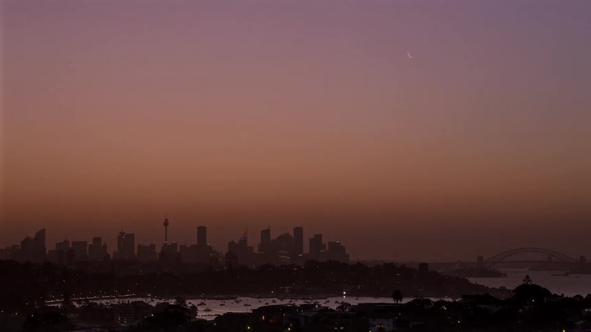 As the glowing sun sets behind the silhouetted skyline the vibrant orange sky fades into deep twilight eventually revealing a delicate crescent moon and thousands of glittering city lights reflected over the darkened harbor