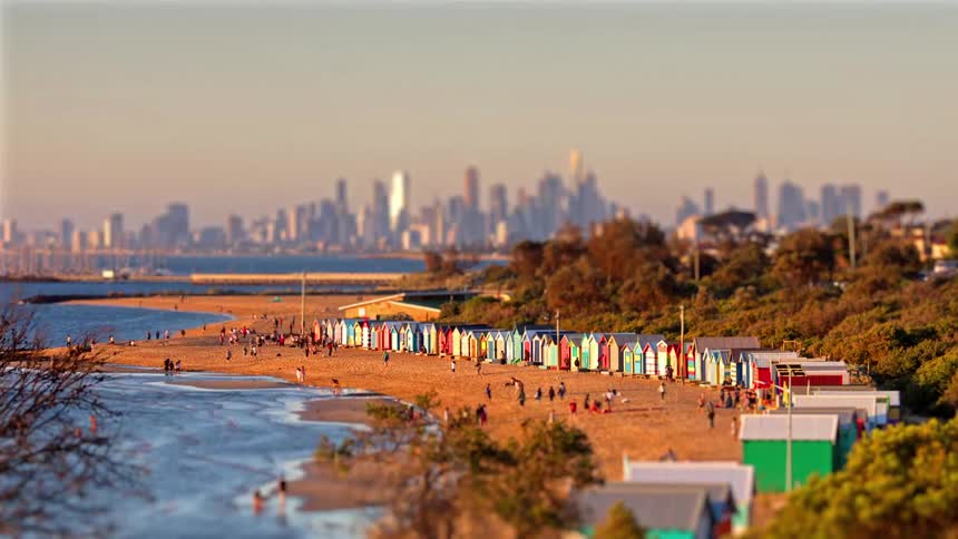 A view of a row of colorful bathing boxes along a sandy beach as the sun sets and city lights begin to glow on the distant skyline