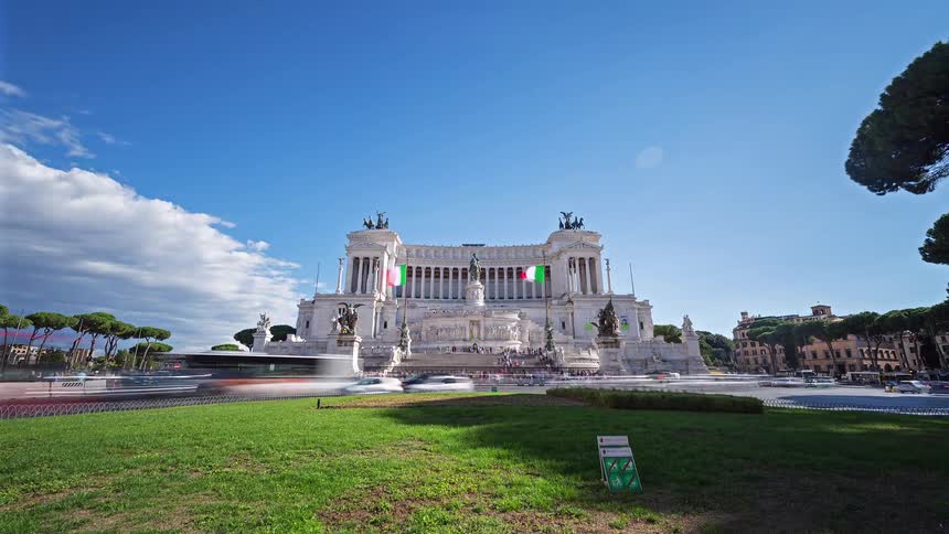 A view from a distance shows a large white building with several statues and two flags on it as many vehicles move along a road in front of it during a sunny day