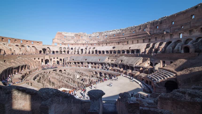 Roman amphitheater with stone seating and visitors spread across the arena under a blue sky