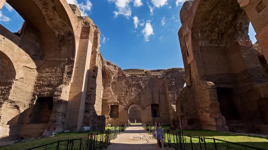 A camera moves through the towering brick ruins of the baths of caracalla under a blue sky with white clouds as tourists walk along gravel paths between ancient stone walls and arched doorways