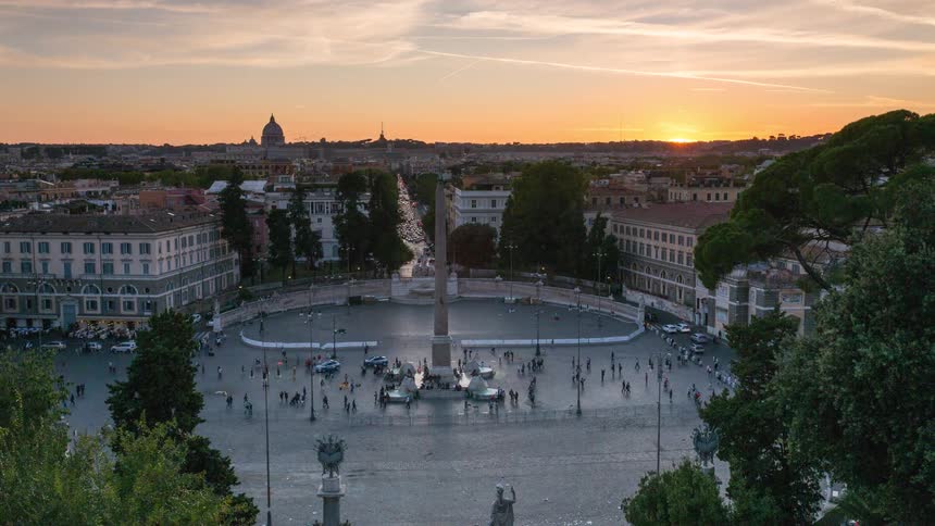 A high angle view captures people moving across a large city square with a tall stone obelisk as the sun sets over the horizon and city lights begin to glow during the transition from day to night