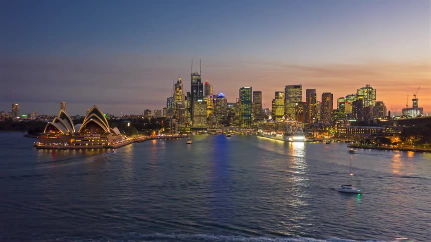 Urban bay at dusk with an opera house moving boats and an illuminated skyline in the background