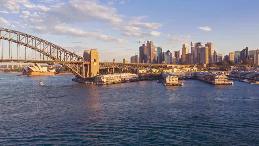 Boats traverse sydney harbor past the iconic bridge and city skyline bathed in golden sunset light