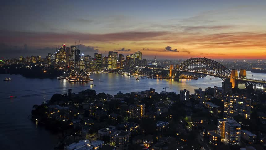 A glowing sunset settles over sydney harbour with boats gliding across the water as the opera house and harbour bridge shine amid the city lights