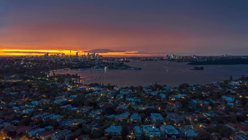 A panoramic aerial view shows a harbor city glowing at dusk with scattered neighborhood lights in the foreground and a vibrant skyline silhouetted against a fiery sunset