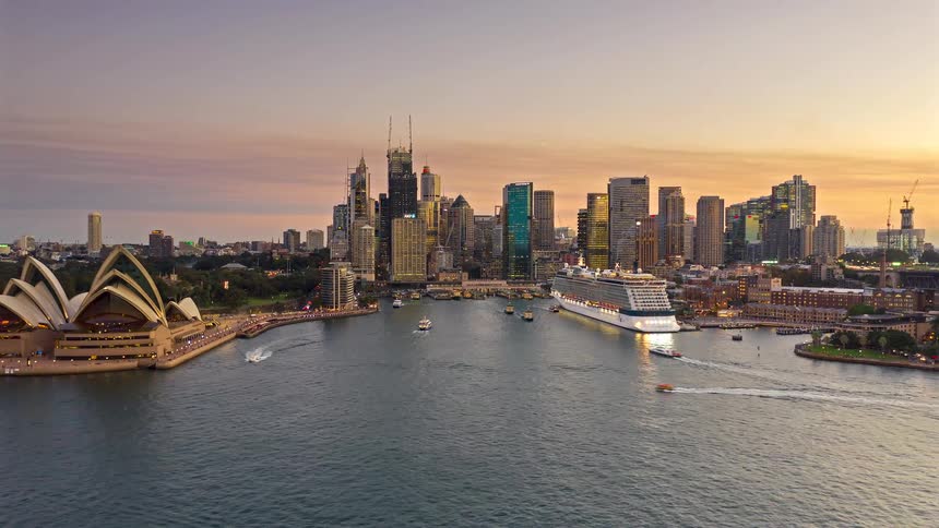 Downtown towers and skyscrapers overlook a cruise ship docked along the coast as boats sail by the sydney opera house