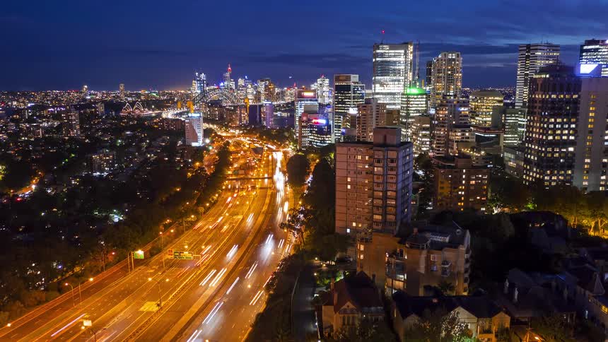 This wide aerial view shows a glowing city skyline at night with buildings illuminated as light trails from traffic blur along a major highway snaking through the city