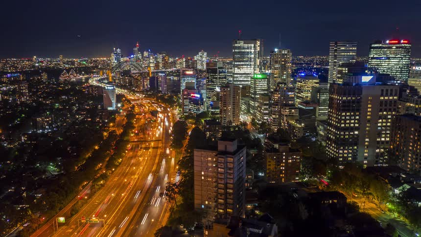 A busy nightlife scene in the city with buildings lit up and the skyline visible as cars speed along a brightly lit highway
