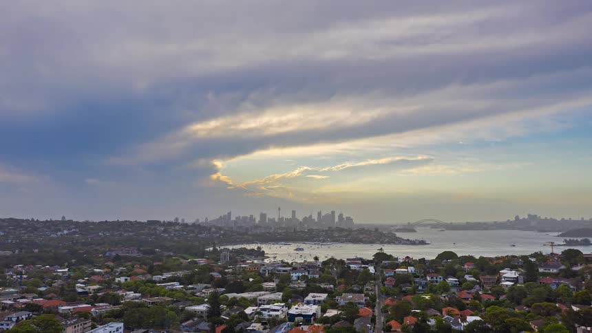 A sweeping cityscape stretches from leafy suburban neighborhoods to a calm lake and distant skyline all beneath dramatic evening clouds glowing with fading light