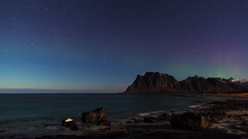 A starfilled night sky stretches over a calm shoreline where dark mountains rise beside a quiet sea under faint bands of auroralike light