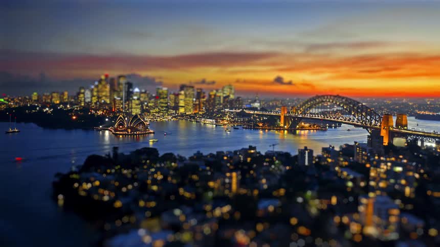 A high angle view shows the city skyline and a large bridge over the harbor as the sun sets behind buildings creating a vibrant orange and yellow sky with silhouettes reflecting on the water
