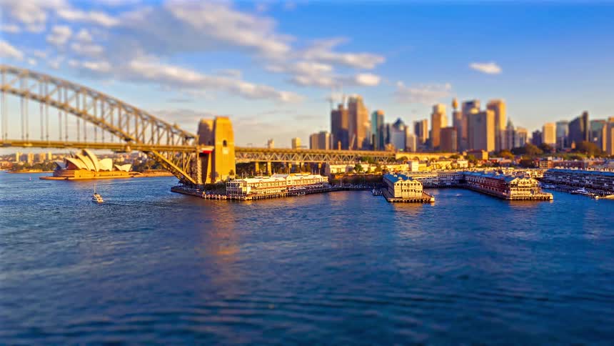 A high angle view shows the city skyline and a large bridge over the harbor as the sun sets behind buildings creating a vibrant orange and yellow sky with silhouettes reflecting on the water
