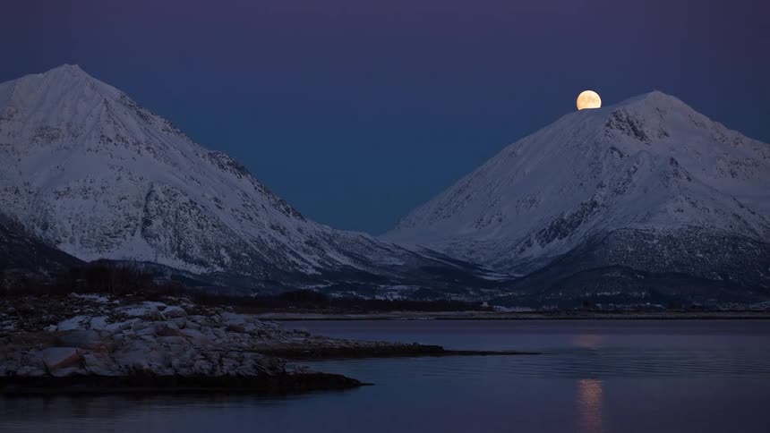 Full moon rising over snow covered mountains along the coast