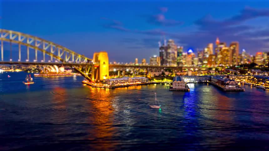 Arch bridge over the bay with boats navigating and an illuminated urban skyline in the background at night