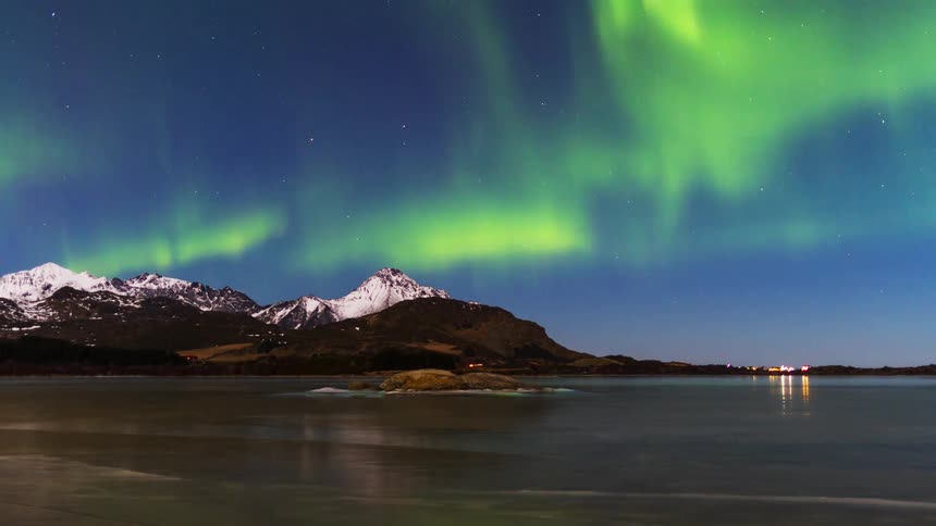 A mountain along the shoreline with green light in the night sky and light shining from a building in the distance