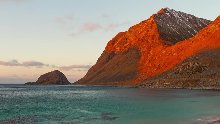 Golden sunset light bathes a steep coastal mountain overlooking a calm turquoise sea