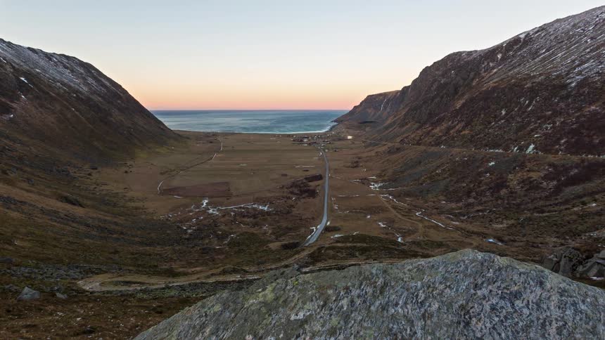 A narrow road winds through a rugged mountain valley toward the ocean bathed in soft light from a calm sunset