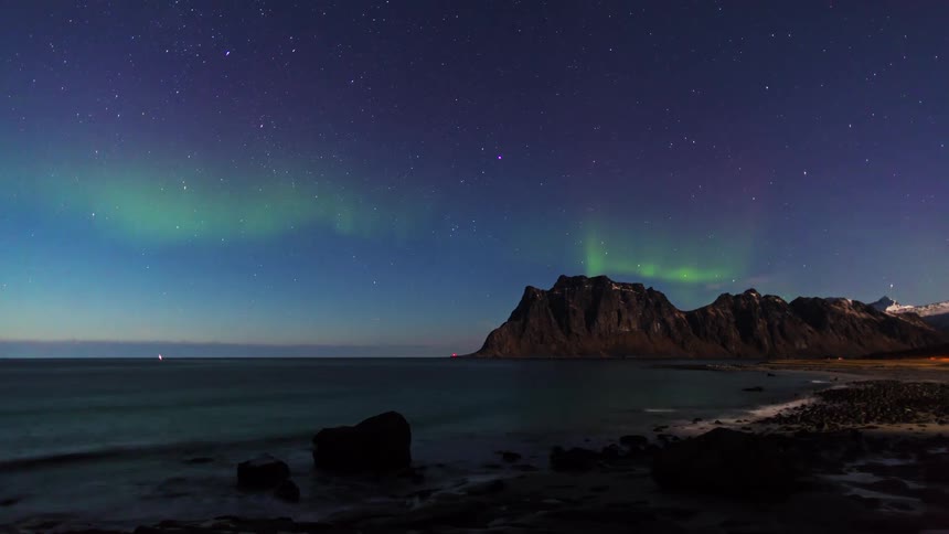 Northern lights over the ocean with a rocky coastline and mountain in the background under a starry night sky
