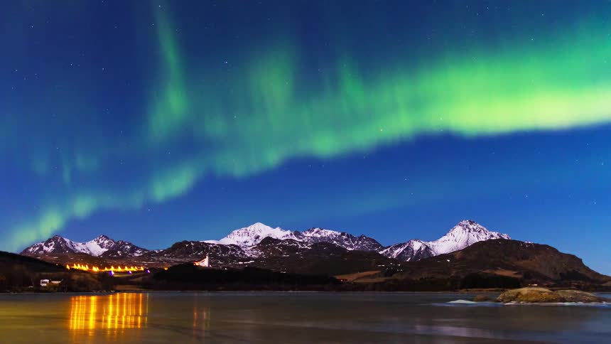 Vibrant green northern lights dance across a starry sky over snow capped mountains and a reflecting lake