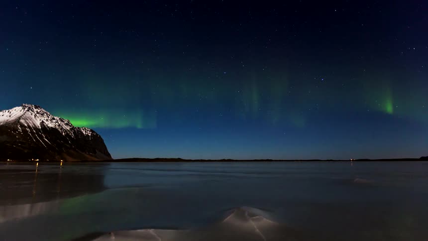 Northern lights glow above a snowy mountain and frozen shoreline under a starry night sky