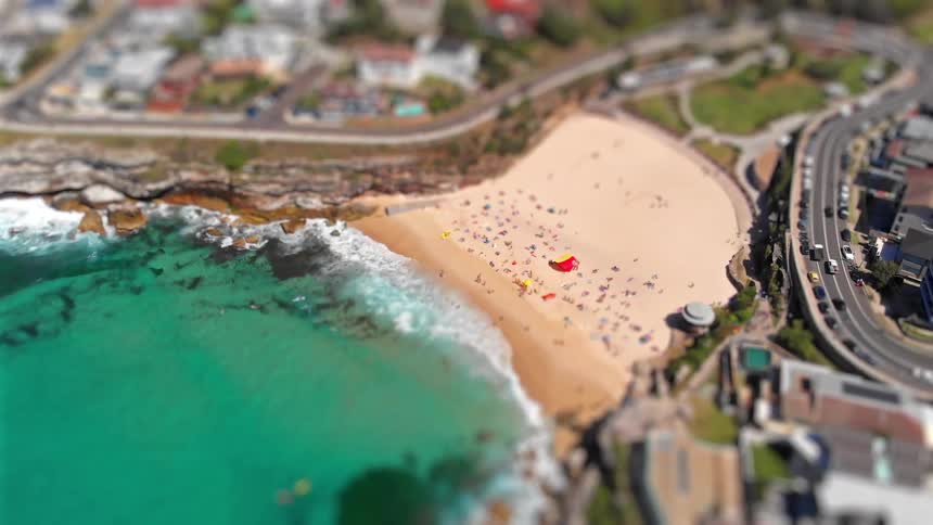 Aerial view of a crowded sandy beach beside turquoise ocean waves and a curving coastal road