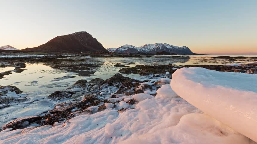 Rocky shoreline with snow and ice in the foreground shallow sea and mountains in the background under early morning light