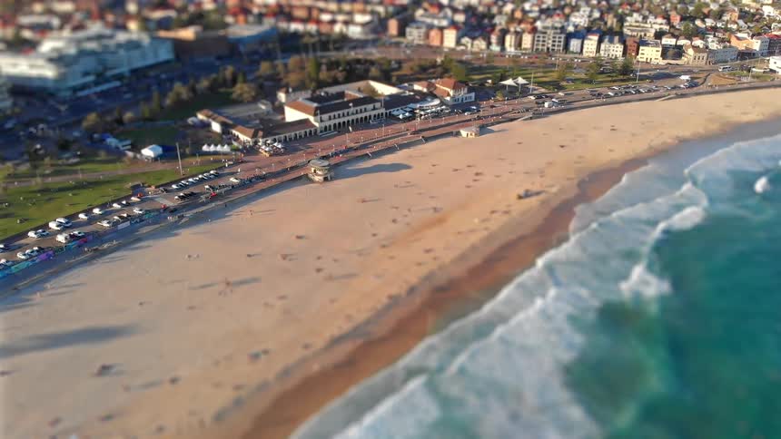 People walk on the beach as waves roll in to shore and traffic moves on the streets of a busy coastal neighborhood