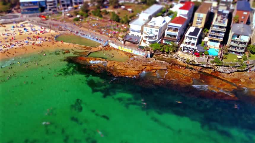 A miniature style aerial view captures a vibrant crowded beach and rocky coastline with turquoise water