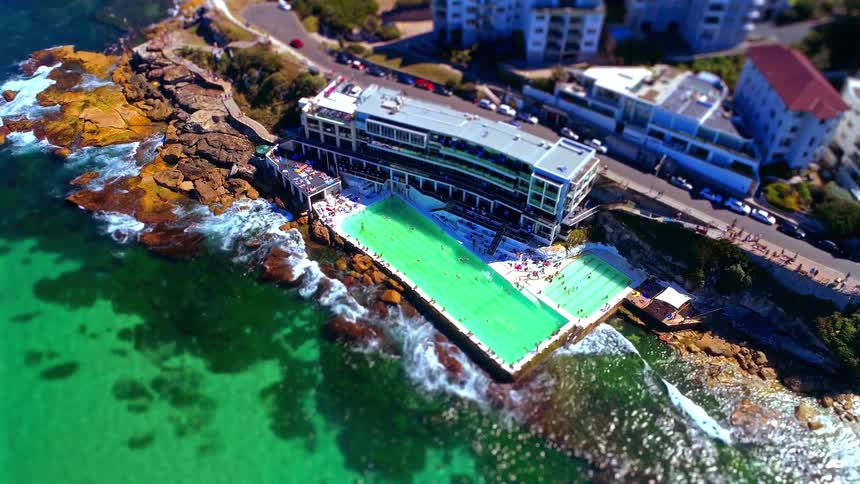 The footage captures a highangle aerial view of a bright green oceanfront swimming pool built directly into a rocky coastline waves from the turquoise sea gently crash against the concrete edges of the pool while people lounge and swim under the bright