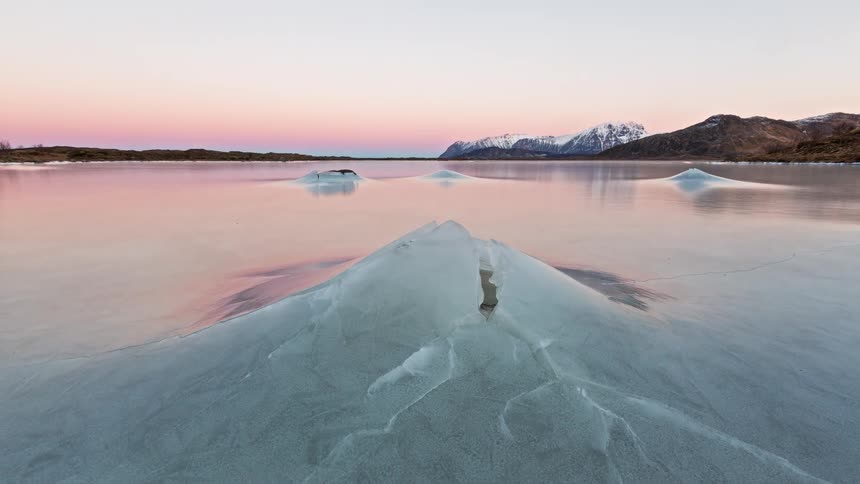 Frozen lake with ice plates in the foreground mountains in the background and a pasteltoned sky