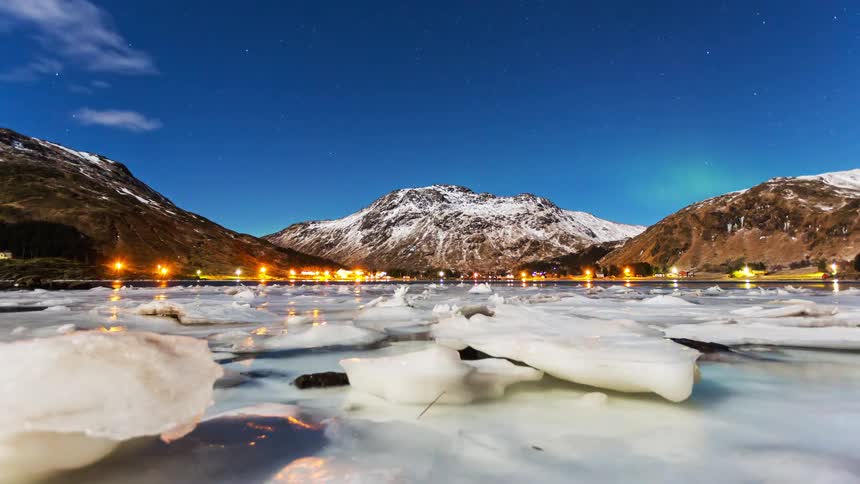 A lake with chunks of ice floating on the surface bright lights shining from buildings along the shoreline and a frozen mountain range behind it at sunrise