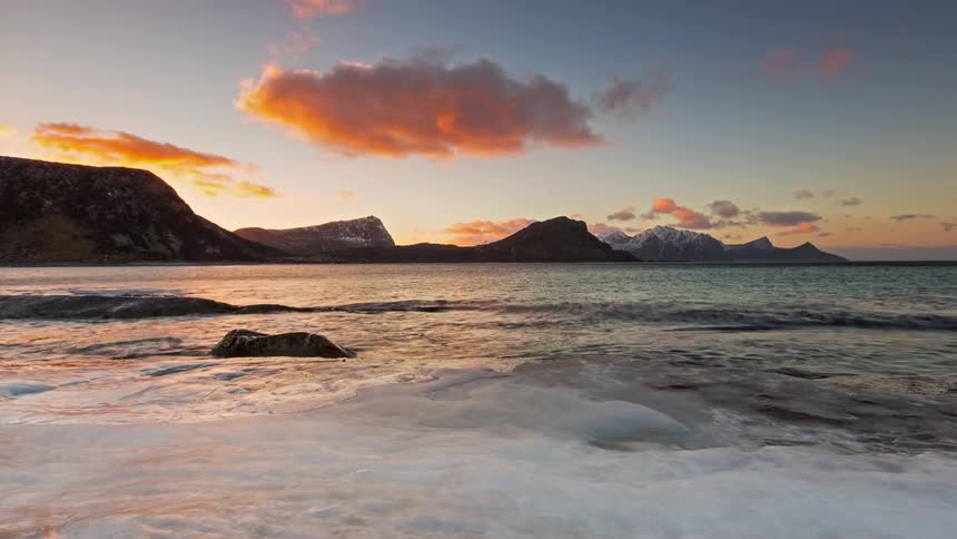 A frozen beach covered with ice as waves hit the shoreline with a mountain range in the background at sunrise