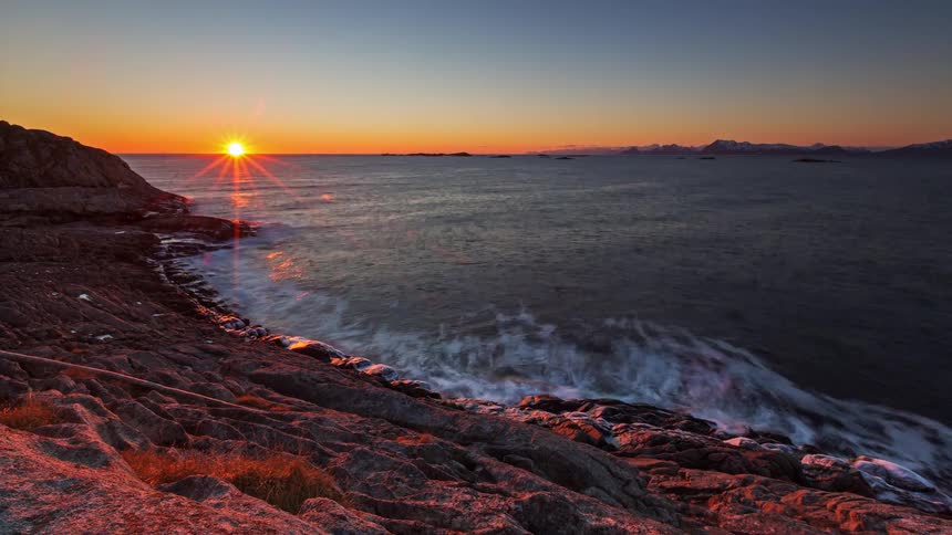 Waves crash against a rocky shoreline as the sun sets over a calm ocean with distant mountains visible under a clear orange and blue sky