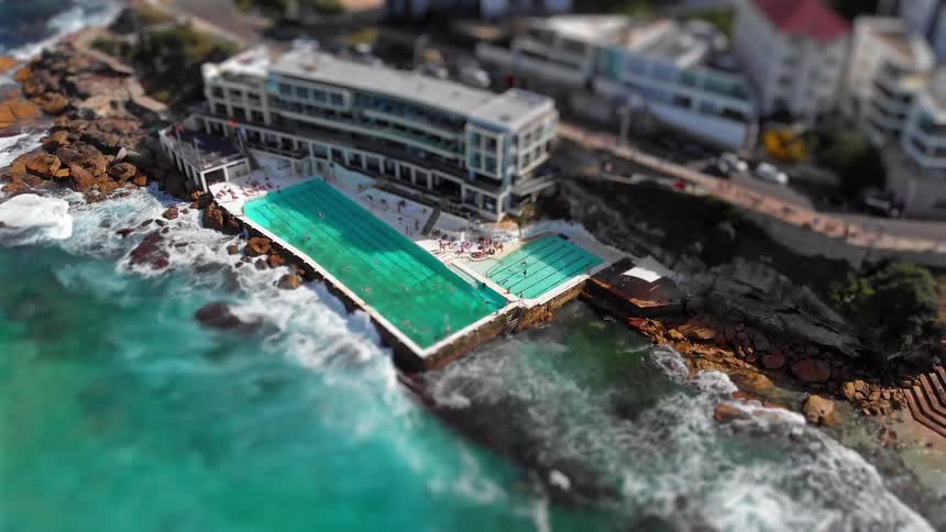 Aerial view of ocean pools with people swimming alongside a rocky shoreline and a waterfront building