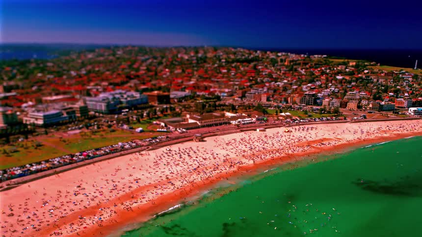 This high angle tilt shift view shows a crowded beach with turquoise water and sandy shores bordered by a dense coastal suburb and ocean pools
