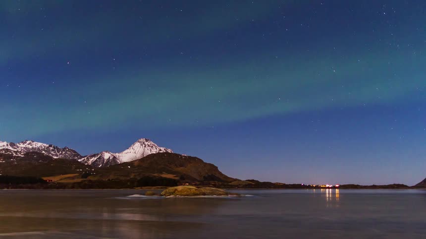 A tranquil night scene shows snowcapped mountains and a still shoreline beneath a starry sky and faint northern lights with a few distant houses glowing softly along the coast