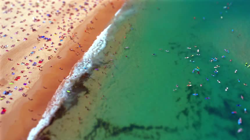 A miniature style aerial view captures a crowded beach and surfers scattered across vibrant turquoise water