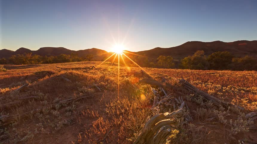 Sun rising on the horizon over a dry field with low vegetation and mountains in the background