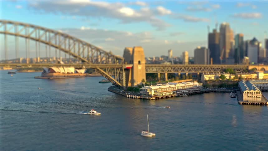 A miniature style aerial view captures boats moving through sydney harbor past the iconic bridge and opera house at sunset