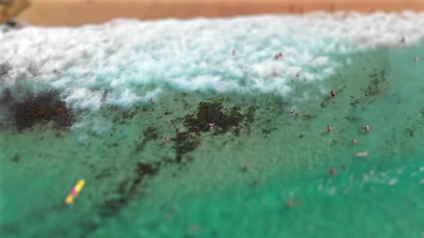 An aerial tilt shift view shows many people swimming in clear turquoise water as white waves crash against a rocky shoreline and sandy beach where a person with a yellow board drifts near the surf