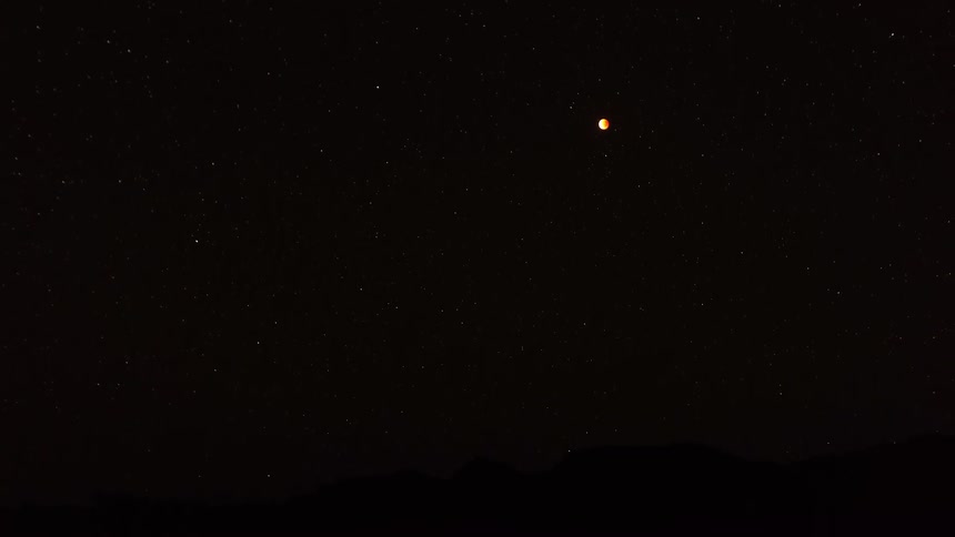A time lapse view shows the bright moon and stars moving across a dark night sky over the silhouettes of desert mountains as the light shifts during the transition to early morning