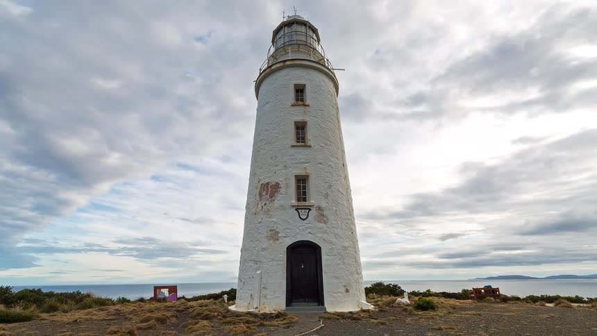 White lighthouse in a coastal area with low vegetation and the ocean in the background under a cloudy sky